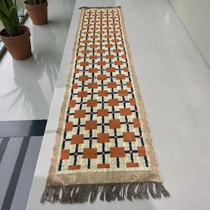 May include: A rectangular runner rug with a geometric pattern of orange squares, black crosses, and pink dots on a cream background. The rug has a tan border and fringe on both ends. The rug is approximately 60 inches long.