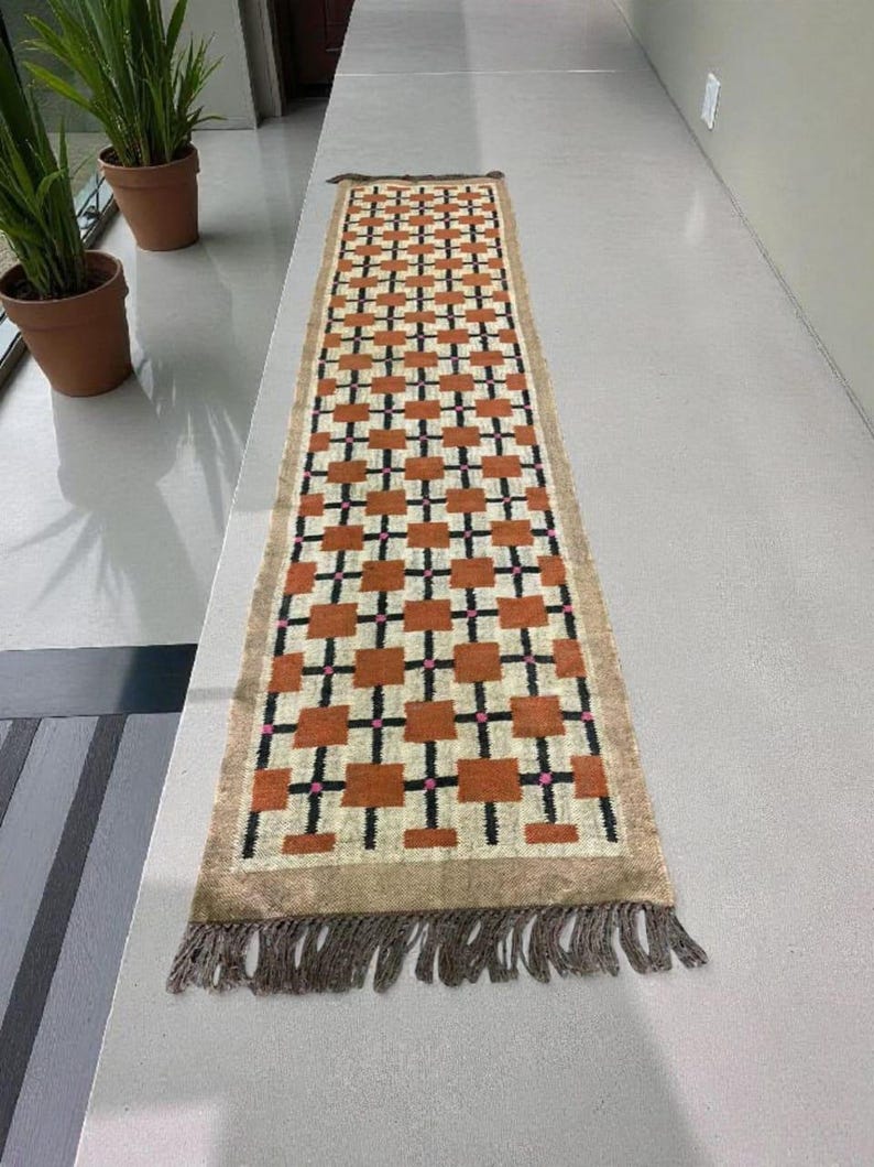 May include: A rectangular runner rug with a geometric pattern of orange squares, black crosses, and pink dots on a cream background. The rug has a tan border and fringe on both ends. The rug is approximately 60 inches long.