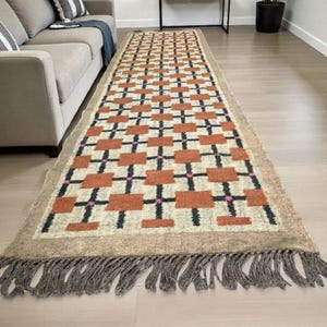 May include: A rectangular area rug with a geometric pattern of rust-colored squares, black crosses, and pink accents on a cream background. The rug has a tan border and fringe.