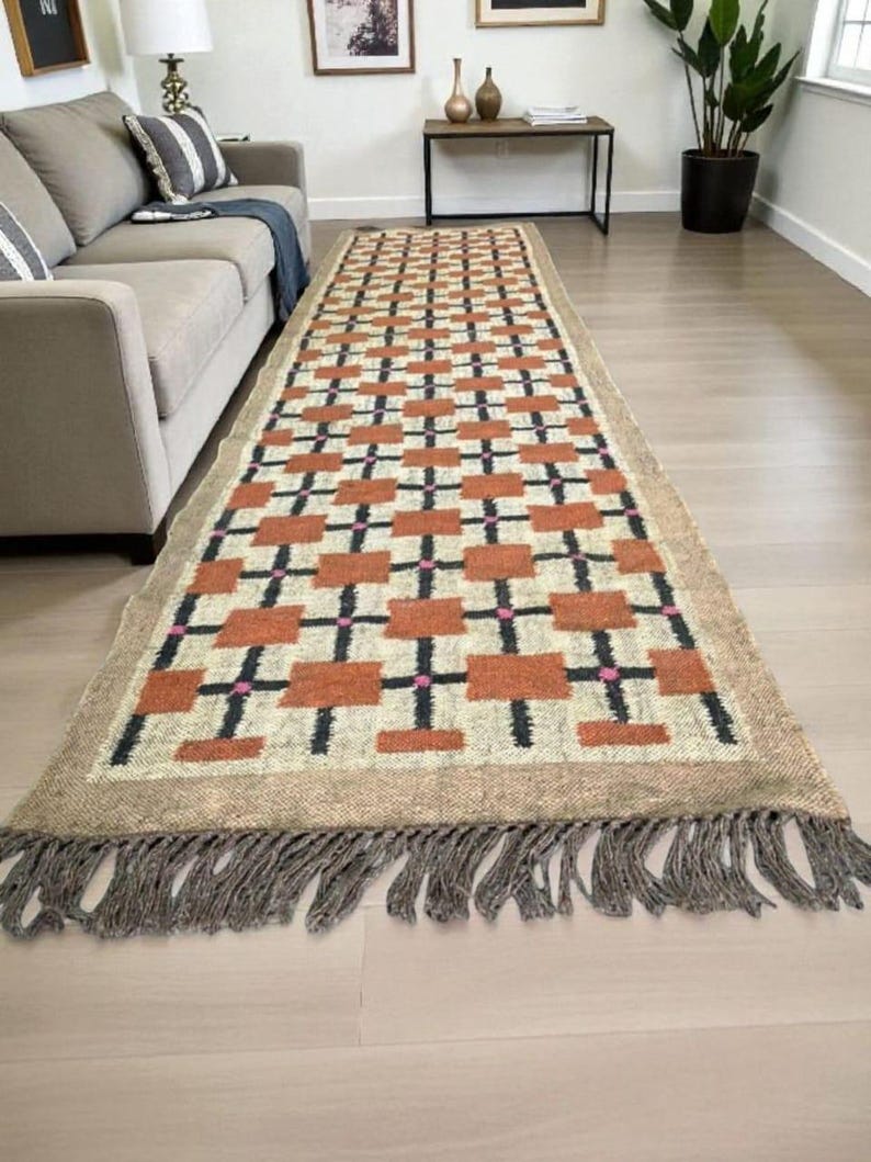 May include: A rectangular area rug with a geometric pattern of rust-colored squares, black crosses, and pink accents on a cream background. The rug has a tan border and fringe.