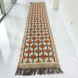 May include: A rectangular hallway rug with a geometric pattern. The rug features a repeating design of brown squares and black crosses on a beige background. Pink accents are visible within the pattern. The rug has a brown fringe on both ends.