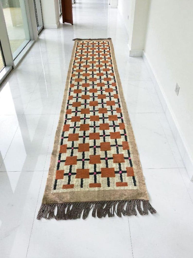 May include: A rectangular hallway rug with a geometric pattern. The rug features a repeating design of brown squares and black crosses on a beige background. Pink accents are visible within the pattern. The rug has a brown fringe on both ends.