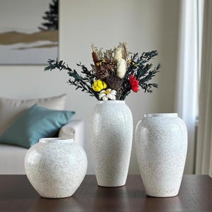 May include: Three speckled white ceramic vases of varying sizes are displayed on a dark wooden surface. The tallest vase holds a bouquet of dried flowers and greenery. The vases have a textured, speckled finish.