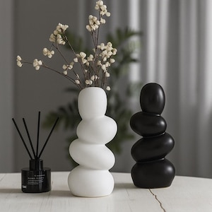 Puede incluir: Un jarrón blanco escultural con un diseño apilado único, que contiene delicadas flores blancas. Junto a él, una escultura de piedra negra apilada. Un frasco difusor negro con cañas está a la izquierda. Los objetos están sobre una superficie blanca.