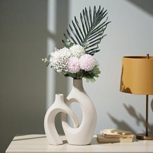 May include: Two cream-coloured ceramic vases with a distinctive, curved design. One vase holds a bouquet of white, pink, and green artificial flowers and foliage. A gold-toned lamp and a stack of books are also visible.