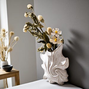 Puede incluir: Un jarr&oacute;n blanco texturizado con un dise&ntilde;o &uacute;nico y arremolinado, lleno de flores secas y vegetaci&oacute;n. El arreglo incluye flores blancas en forma de margarita y cabezas de semillas esponjosas de color canela. El jarr&oacute;n est&aacute; colocado sobre una superficie blanca contra una pared gris.