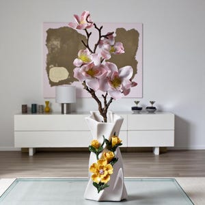May include: A white ceramic vase with a twisted design, adorned with yellow and green floral accents. The vase holds a branch of pink and yellow artificial flowers. The arrangement is set against a modern white cabinet and a pink abstract painting.