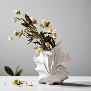 Puede incluir: Un jarr&oacute;n de cer&aacute;mica blanco en forma de concha con una superficie texturizada, que contiene flores secas y vegetaci&oacute;n. El jarr&oacute;n tiene un dise&ntilde;o en espiral y est&aacute; lleno de flores blancas y amarillas. La est&eacute;tica general es natural y decorativa.