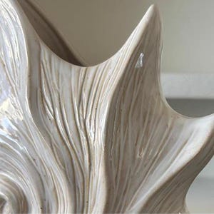 Puede incluir: Primer plano de una pieza decorativa de cer&aacute;mica color crema con un dise&ntilde;o texturizado en forma de cuerno. La superficie tiene ranuras verticales y un acabado brillante, creando una est&eacute;tica natural y org&aacute;nica. La pieza tiene una forma suave y curvada.