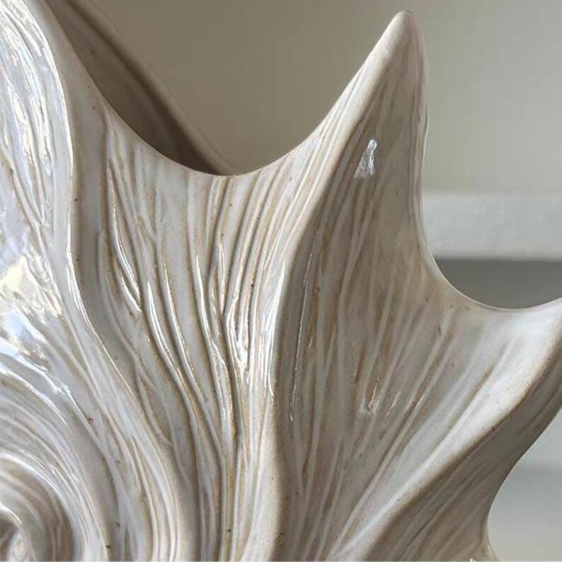 Puede incluir: Primer plano de una pieza decorativa de cer&aacute;mica color crema con un dise&ntilde;o texturizado en forma de cuerno. La superficie tiene ranuras verticales y un acabado brillante, creando una est&eacute;tica natural y org&aacute;nica. La pieza tiene una forma suave y curvada.