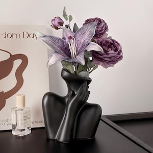 Puede incluir: Un jarrón de cerámica negra con forma de cabeza y hombros, con las manos cubriendo la cara. El jarrón contiene flores y vegetación moradas. A la izquierda del jarrón hay un frasco de perfume.