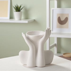 Könnte beinhalten: Eine weiße Keramikvase in Form von Händen, die eine Schale halten. Die Vase hat eine glatte, matte Oberfläche und ist für kleine Pflanzen oder Dekorationsgegenstände konzipiert. Die Hände sind detailliert und erzeugen einen einzigartigen, künstlerischen Look.
