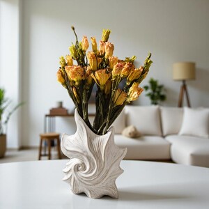 Puede incluir: Un jarr&oacute;n blanco en forma de concha marina contiene un ramo de flores secas de color naranja y amarillo. El jarr&oacute;n tiene una superficie texturizada y un dise&ntilde;o en espiral. Las flores tienen tallos verdes y est&aacute;n dispuestas de forma natural. El jarr&oacute;n est&aacute; sobre una mesa blanca.