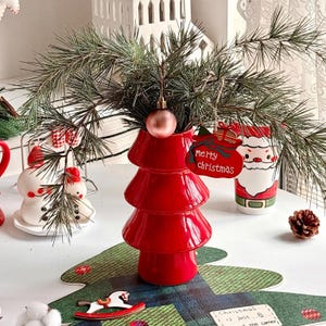 Puede incluir: Un jarrón rojo de cerámica con forma de árbol de Navidad contiene ramas de pino, una bola rosa y una etiqueta "Merry Christmas". También se ven una taza de Papá Noel y figuras de muñecos de nieve, creando una escena festiva.