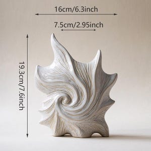 Puede incluir: Un jarr&oacute;n decorativo de cer&aacute;mica blanca con un dise&ntilde;o &uacute;nico y arremolinado. El jarr&oacute;n mide 19,3 cm de alto, 16 cm de ancho y 7,5 cm de profundidad. La superficie texturizada presenta un patr&oacute;n en espiral y bordes puntiagudos.