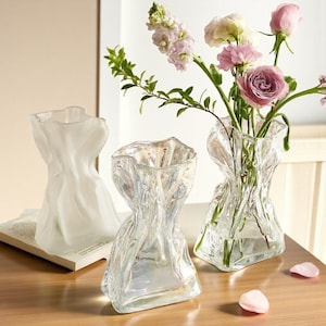 May include: Three clear glass vases with a textured, folded design. One vase is filled with pink and purple flowers. The vases are on a wooden surface, with a book and scattered petals nearby.