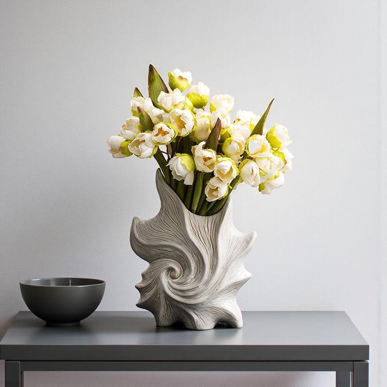 Puede incluir: Un jarr&oacute;n decorativo con un dise&ntilde;o arremolinado &uacute;nico contiene un ramo de flores blancas y amarillas con hojas verdes. Un peque&ntilde;o cuenco gris oscuro est&aacute; a la izquierda del jarr&oacute;n. El jarr&oacute;n y el cuenco est&aacute;n sobre una mesa gris.