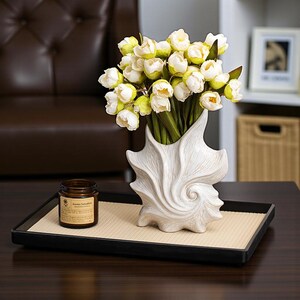Puede incluir: Un jarr&oacute;n blanco en forma de concha con tulipanes blancos y amarillos sobre una bandeja negra con una esterilla beige. Tambi&eacute;n hay un peque&ntilde;o frasco marr&oacute;n con una etiqueta en la bandeja.