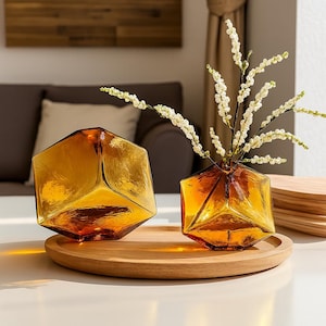 Puede incluir: Dos jarrones geométricos de vidrio ámbar, uno con flores blancas, sobre una bandeja redonda de madera. Los jarrones tienen una superficie texturizada y un diseño angular. El conjunto se encuentra sobre un fondo neutro. Altura de los jarrones: aprox. 15 cm.