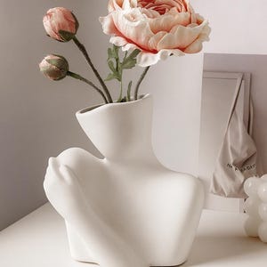 Könnte beinhalten: Eine weiße Keramikvase in Form eines Torsos hält Kunstblumen. Die Anordnung umfasst rosa Knospen und eine große, pfirsichfarbene Pfingstrose. Die Vase hat eine glatte, matte Oberfläche und steht auf einer weißen Oberfläche.