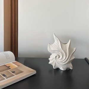 Puede incluir: Un jarr&oacute;n de cer&aacute;mica abstracto de color crema con un dise&ntilde;o en espiral y forma de hoja. El jarr&oacute;n tiene una superficie texturizada y est&aacute; colocado sobre una mesa negra. Es un elemento decorativo para el hogar.