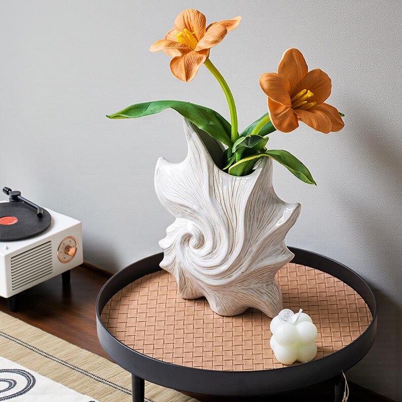 Puede incluir: Un jarr&oacute;n de cer&aacute;mica blanca texturizada con un dise&ntilde;o en espiral contiene flores naranjas con hojas verdes. El jarr&oacute;n est&aacute; sobre una mesa redonda negra con una superficie tejida. Tambi&eacute;n hay una peque&ntilde;a vela blanca sobre la mesa.