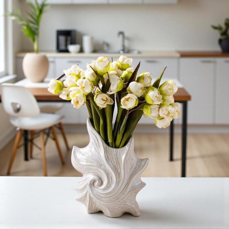 Puede incluir: Un jarr&oacute;n de cer&aacute;mica blanca con un dise&ntilde;o en espiral contiene un ramo de flores blancas y amarillas. El jarr&oacute;n est&aacute; sobre una superficie blanca, con una cocina y un comedor al fondo.