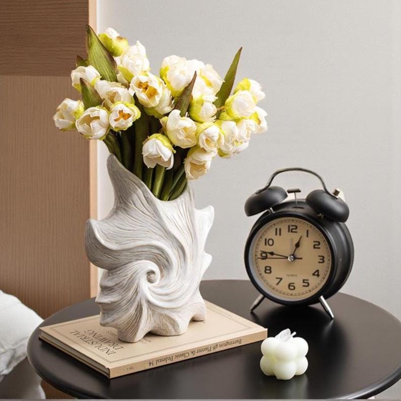 Puede incluir: Un jarr&oacute;n blanco en forma de concha contiene un ramo de flores blancas y amarillas. Un despertador negro y una peque&ntilde;a vela blanca est&aacute;n sobre una mesa negra. Un libro con el texto "INDUSTRIAL DRAWING" est&aacute; debajo del jarr&oacute;n.