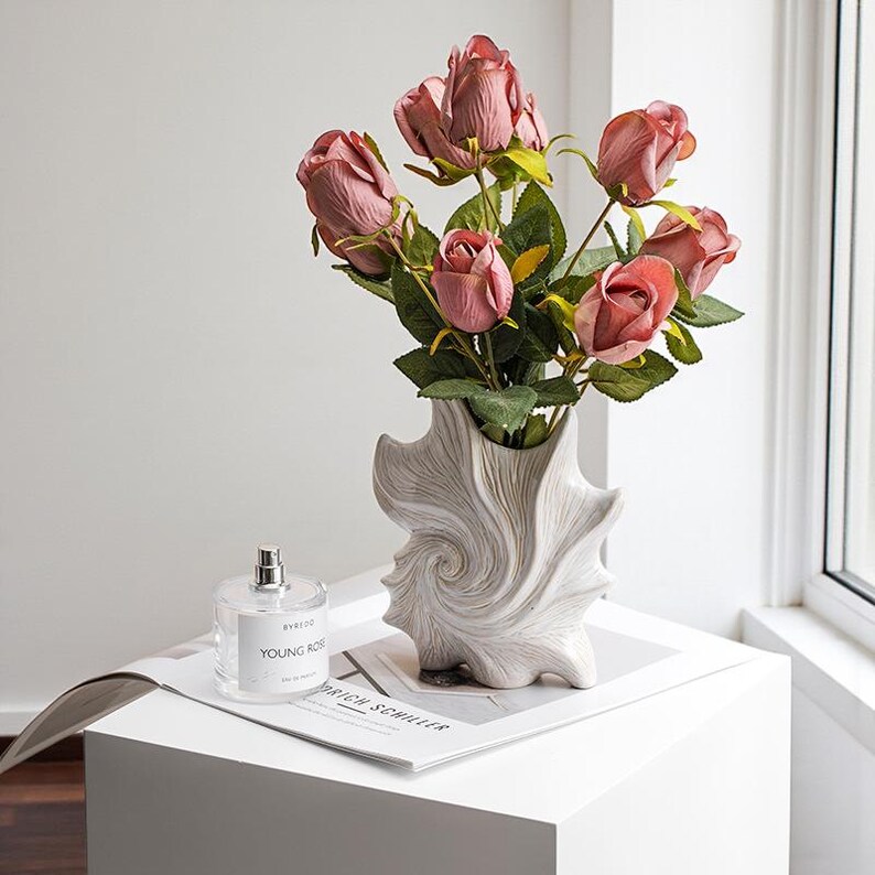 Puede incluir: Un arreglo decorativo de rosas rosas en un jarr&oacute;n &uacute;nico, texturizado y blanquecino. El jarr&oacute;n tiene un dise&ntilde;o en espiral. Un frasco de perfume y una revista est&aacute;n sobre una superficie blanca.