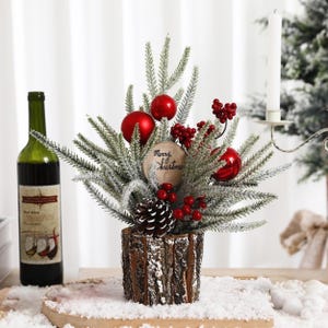 Op de afbeelding: Een feestelijk kerststuk met bevroren dennentakken, rode ballen en bessen in een rustieke houten pot. Een houten schijf met "Merry Christmas" is inbegrepen. Een fles wijn staat links.
