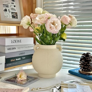 Könnte beinhalten: Eine cremefarbene, strukturierte Keramikvase mit zwei Griffen hält einen Strauß rosa und weißer Blumen. Die Vase steht auf einer weißen Oberfläche, im Hintergrund sind Bücher und ein gerahmtes Bild zu sehen. Eine Perlenkette und ein Tannenzapfen sind ebenfalls sichtbar.
