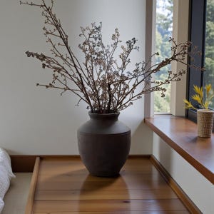 Puede incluir: Un jarrón de cerámica marrón oscuro lleno de ramas y bayas secas. El jarrón está sobre una superficie de madera, cerca de una ventana con vistas a los árboles. Un pequeño jarrón tejido con flores amarillas está en el alféizar.