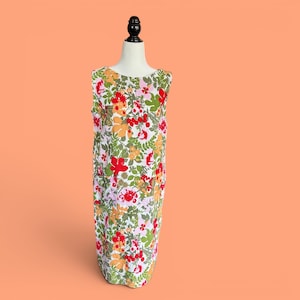 May include: A sleeveless floral dress featuring red, orange, pink, and green flowers on a white base. The dress is displayed on a mannequin against a peach background. The dress has a simple, straight cut. The dress is a summer garment.