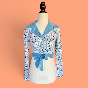 Könnte beinhalten: Ein Oberteil im Vintage-Stil mit langen Ärmeln, hellblauem Kragen und Manschetten. Das Oberteil hat eine weiße Basis mit einem Muster aus blauen, lila und schwarzen Punkten. Es hat eine Schleife an der Taille und ist vorne mit rosa Knöpfen geknöpft.