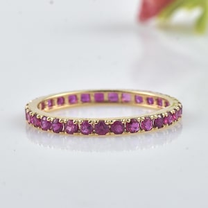 May include: A gold band ring set with a continuous row of vibrant, round-cut rubies. The ring's setting is a warm, yellow gold, and the rubies are a deep, rich pink-red color. The ring is displayed on a white surface.