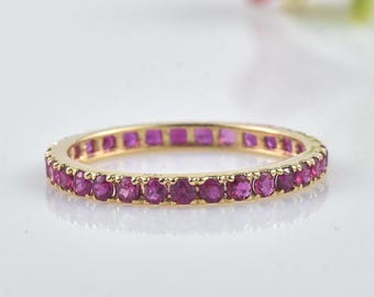 May include: A gold band ring set with a continuous row of vibrant, round-cut rubies. The ring's setting is a warm, yellow gold, and the rubies are a deep, rich pink-red color. The ring is displayed on a white surface.