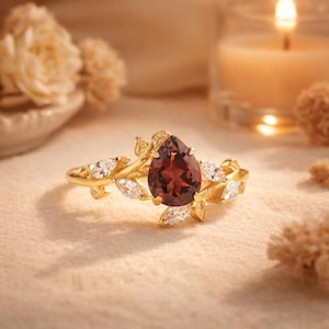 May include: A gold ring with a deep red, pear-shaped gemstone at its centre. The ring is decorated with leaf and marquise-cut white gemstones. The ring is set against a soft, neutral background, with a lit candle in the background.