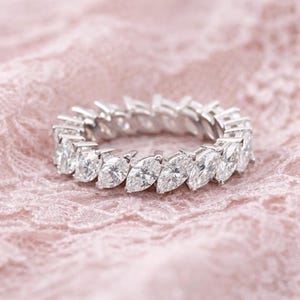 Peut inclure: Une bague en argent ornée d'une rangée continue de diamants taille marquise. La bague est posée sur une surface en dentelle rose, créant un contraste entre l'éclat des diamants et le fond doux. Les diamants sont clairs et réfléchissent la lumière.