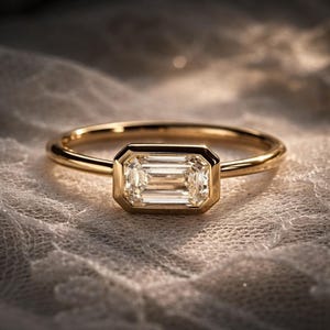 Op de afbeelding: Een gouden ring met een smaragdgeslepen diamant. De diamant is gezet in een bezel-zetting en de band is dun en gepolijst. De ring wordt gepresenteerd op een getextureerd, gebroken witte ondergrond.