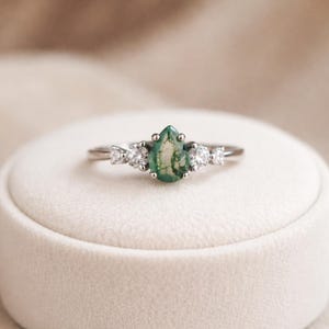 May include: A silver ring featuring a pear-shaped, moss green gemstone as the centerpiece, flanked by smaller, clear gemstones. The ring is set on a white surface, showcasing its delicate design and sparkle.