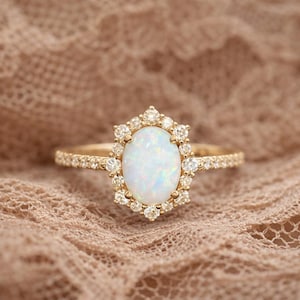 Puede incluir: Un anillo de oro con una piedra de ópalo ovalada, rodeada por un halo de pequeños diamantes redondos. La banda del anillo también está adornada con pequeños diamantes. El anillo se presenta sobre un fondo neutro.