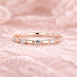 Pode incluir: Anel de banda em ouro rosa com pedras claras retangulares e baguette alternadas. O anel está sobre um fundo rosa suave e texturizado. Uma peça de joalharia delicada.