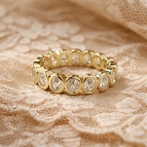 May include: A gold ring featuring a continuous band of oval, clear gemstones. The ring is set against a beige lace background, creating a contrast between the shiny metal and the delicate texture. The gemstones are evenly spaced around the ring.