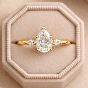 Könnte beinhalten: Ein goldener Verlobungsring mit einem birnenförmigen Diamanten als Mittelstein und zwei kleineren Seitensteinen. Der Ring ist in einem goldenen Band gefasst und wird in einer beigen Ringschatulle präsentiert.