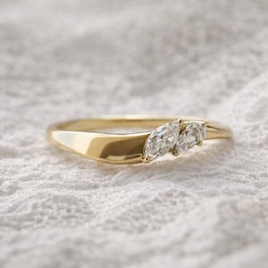Op de afbeelding: Een gouden ring met twee marquise-geslepen diamanten. De ring heeft een gladde, gepolijste band die zachtjes buigt. De diamanten zijn dicht bij elkaar geplaatst, waardoor een sprankelend brandpunt ontstaat. De ring wordt gepresenteerd op een wit, getextureerd oppervlak.