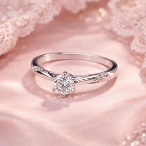 Puede incluir: Un anillo de compromiso plateado con un diamante redondo de talla brillante como piedra central. El anillo presenta una banda delicada con pequeñas piedras de acento a cada lado de la piedra central. Sobre un fondo rosa suave.