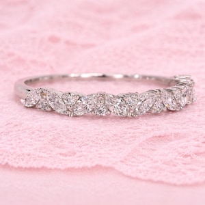 May include: A silver ring featuring a band of marquise-cut diamonds. The diamonds are set closely together, creating a continuous sparkle. The ring is displayed on a pink, textured surface, highlighting its brilliance.