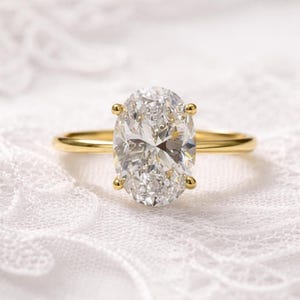 Peut inclure: Une bague de fiançailles en or avec un grand diamant de taille ovale. Le diamant clair et étincelant est serti de quatre griffes sur un fin anneau en or poli. La bague est présentée sur une surface blanche texturée.
