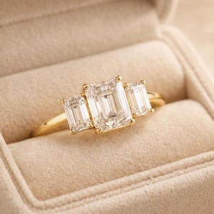 May include: A gold ring featuring three emerald-cut diamonds. The central diamond is the largest, flanked by two smaller stones. The ring is presented in a beige ring box, showcasing the jewellery.