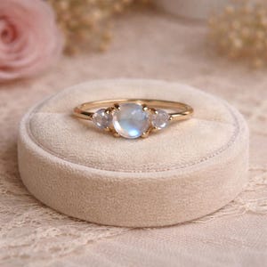 Puede incluir: Un anillo de oro con tres piedras lunares. La piedra central, redonda, es más grande, flanqueada por dos piedras redondas más pequeñas. El anillo descansa sobre una caja de anillo de terciopelo beige, con un fondo floral suave.